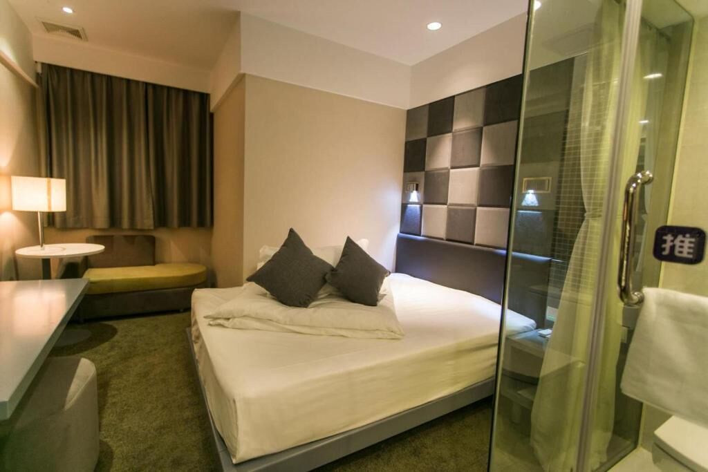 A hotel room with a bed, bedside table, and a glass-enclosed shower area.