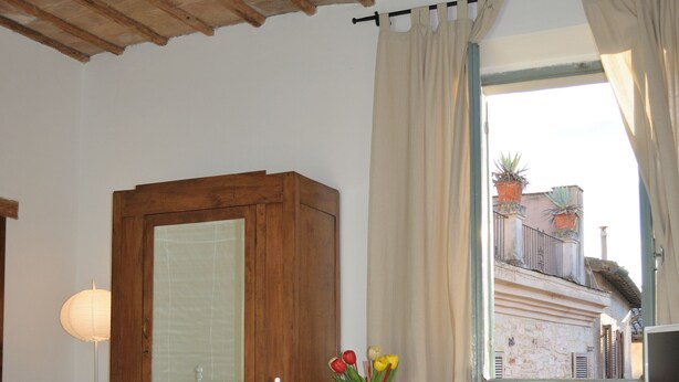 A room with a bed, a small table with a vase of flowers, a chair, and a window overlooking a building.