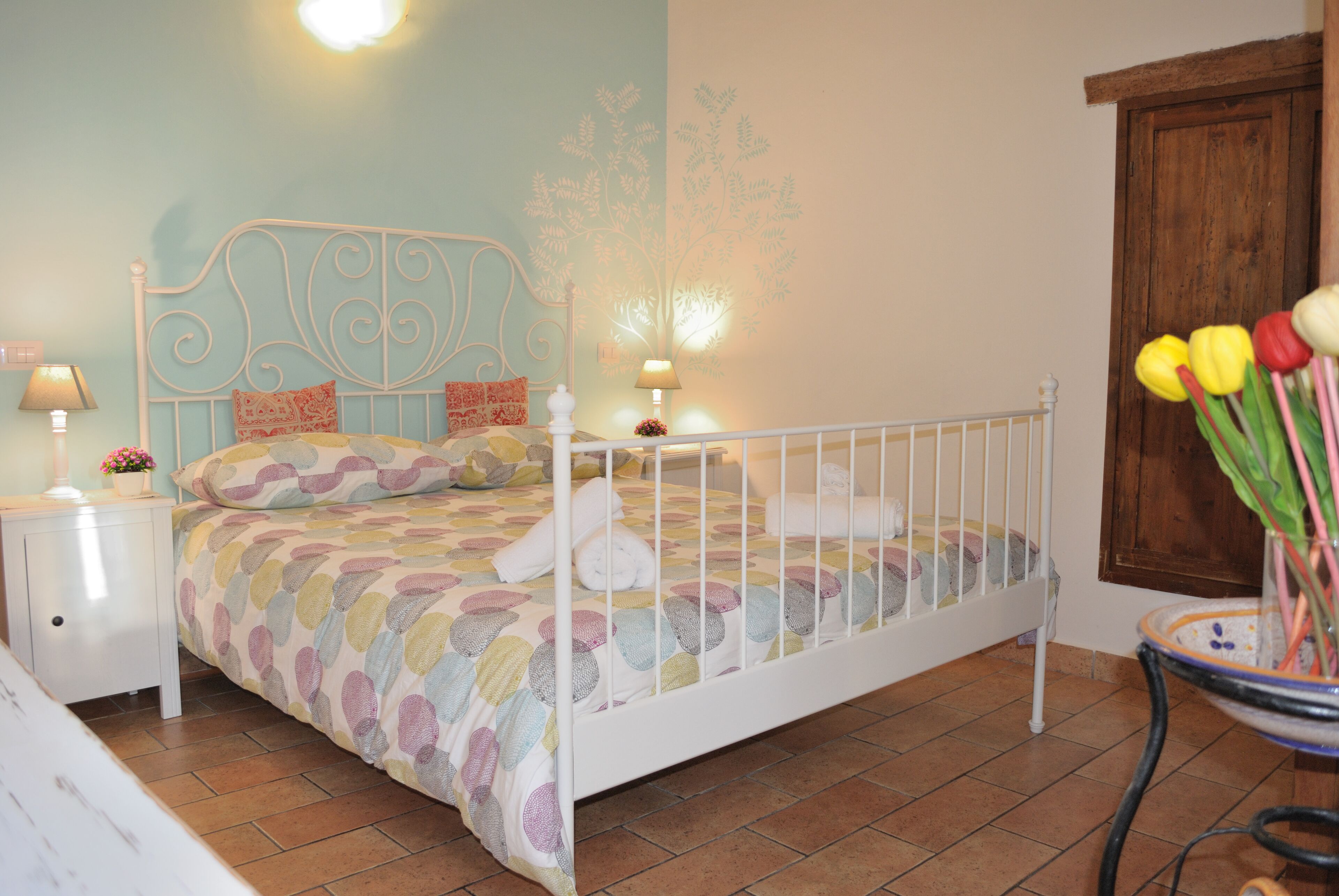 A bedroom with a white metal bed frame, a colorful bedspread, a nightstand with a lamp, and a vase with flowers.