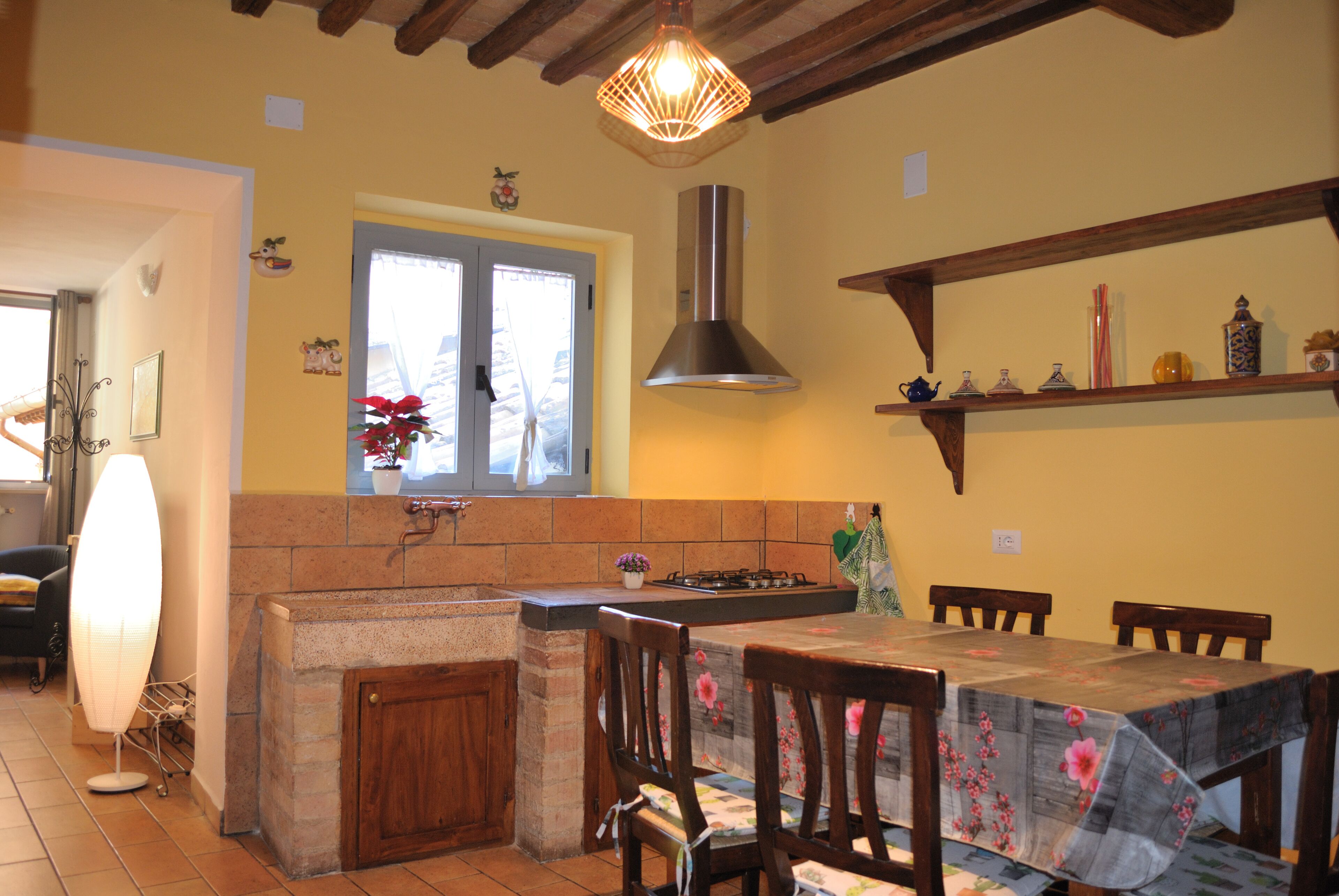 A kitchen with a central island, wooden chairs, a dining table with a floral tablecloth, a hanging light fixture, and a shelf with decorative items.