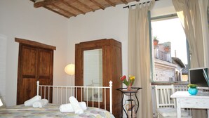 A bedroom with a bed, a wooden wardrobe, a small table with a vase of flowers, a chair, and a window with a view of a balcony.