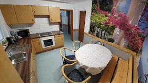 A compact kitchen with wooden cabinets, a microwave, an oven, and a sink. There is a round table with four chairs and a floral tablecloth. A painting of pink flowers is on the wall. - Zum Odenwald (Gorxheimertal)