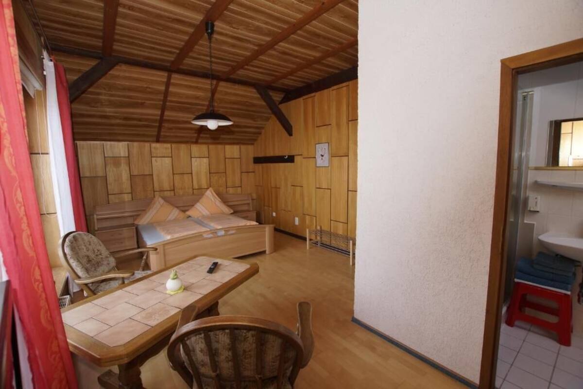 A cozy room with a wooden ceiling, a bed with pillows, a small table with a lamp, and a chair.