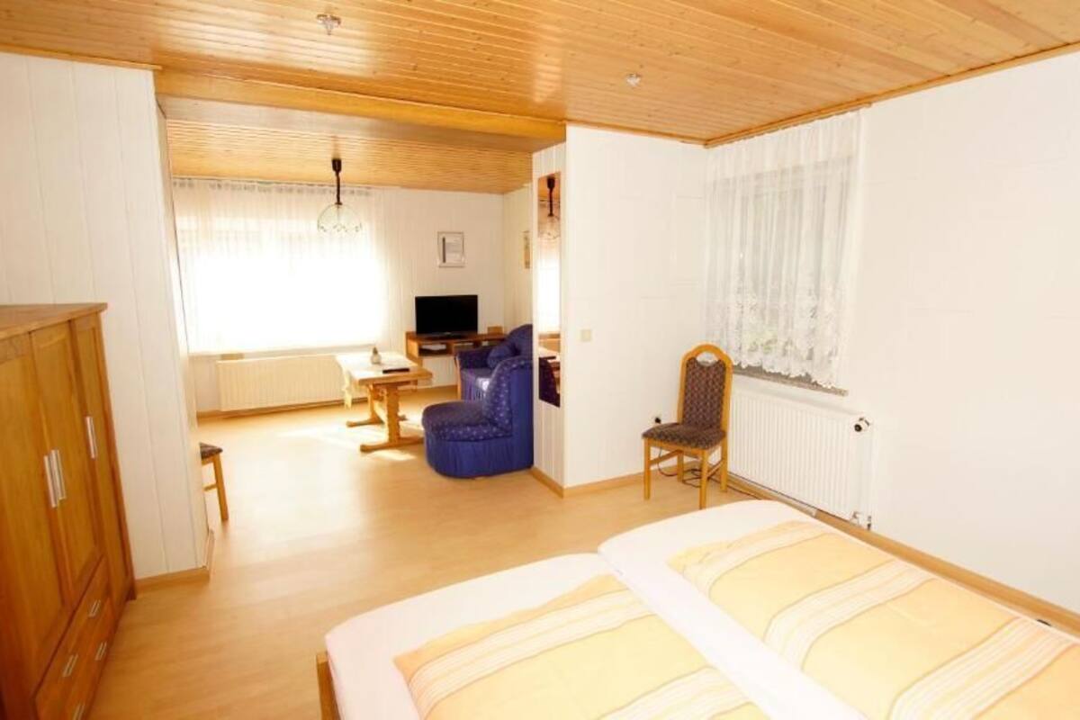 A room with a bed, wooden wardrobe, a small table, a blue chair, and a television.