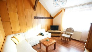 A cozy living room with a white sofa, a wooden coffee table, and a wooden chair. A television is mounted on the wall, and a window provides natural light. - Nichtraucher-Pension Zum Froschkönig (Lübbenau/Spreewald)