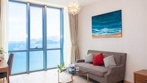 A modern living room with a large window, a sofa with red and grey cushions, a coffee table, and a painting of a beach scene. - OPOX Panorama (Nha Trang)