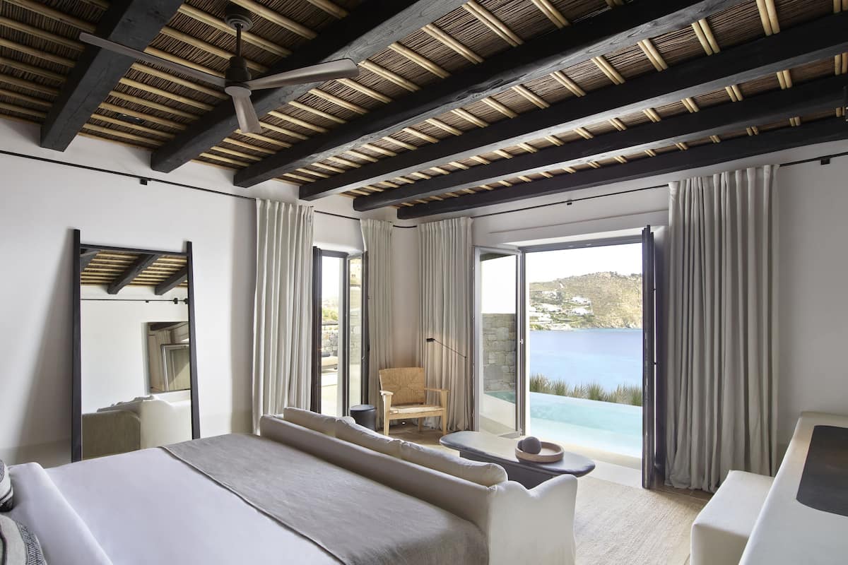 A modern hotel room with a large bed, a seating area with a chair, and a view of the sea through a sliding glass door.