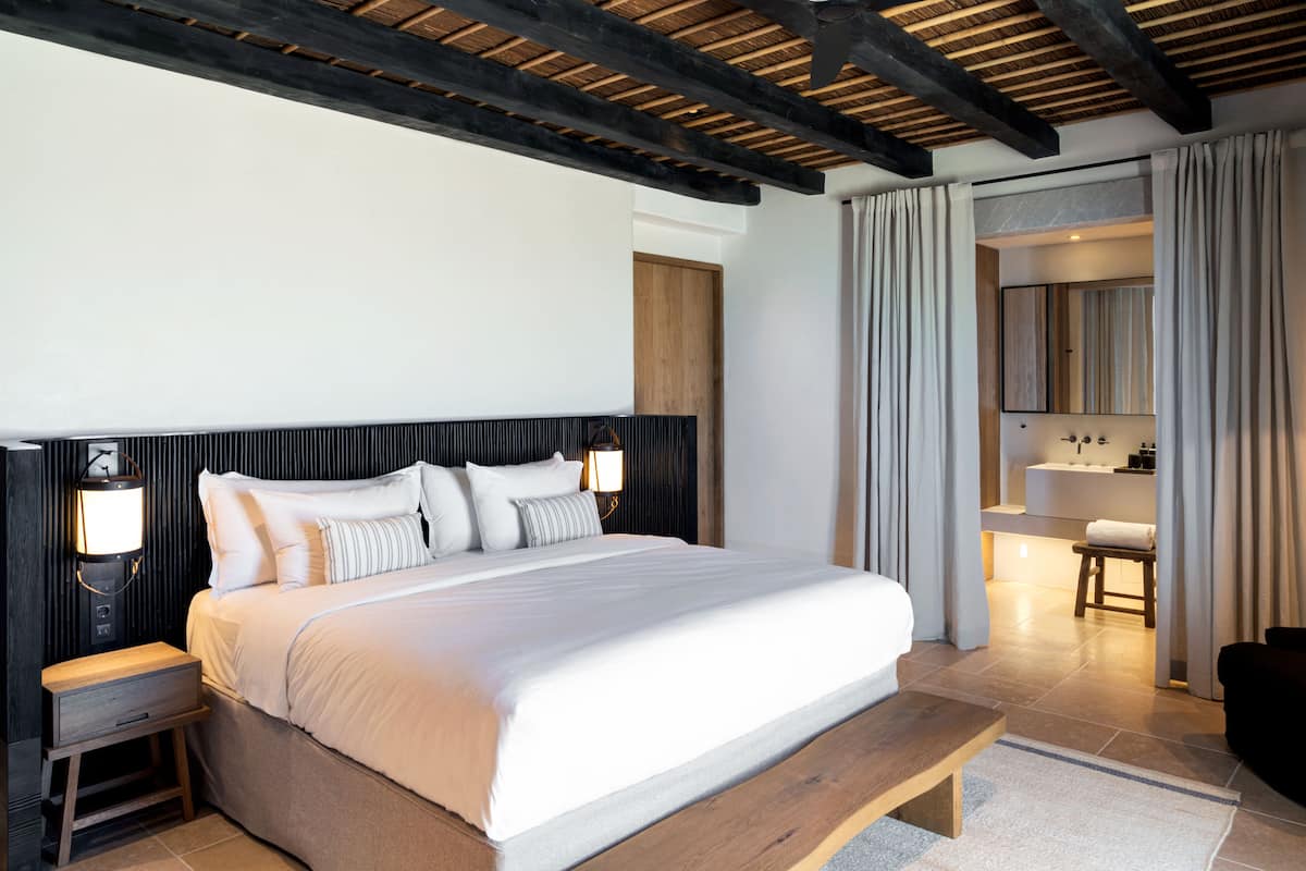 A modern hotel room with a large bed, wooden headboard, bedside tables, and a nightstand. There is a bathroom visible in the background with a bathtub and a mirror.