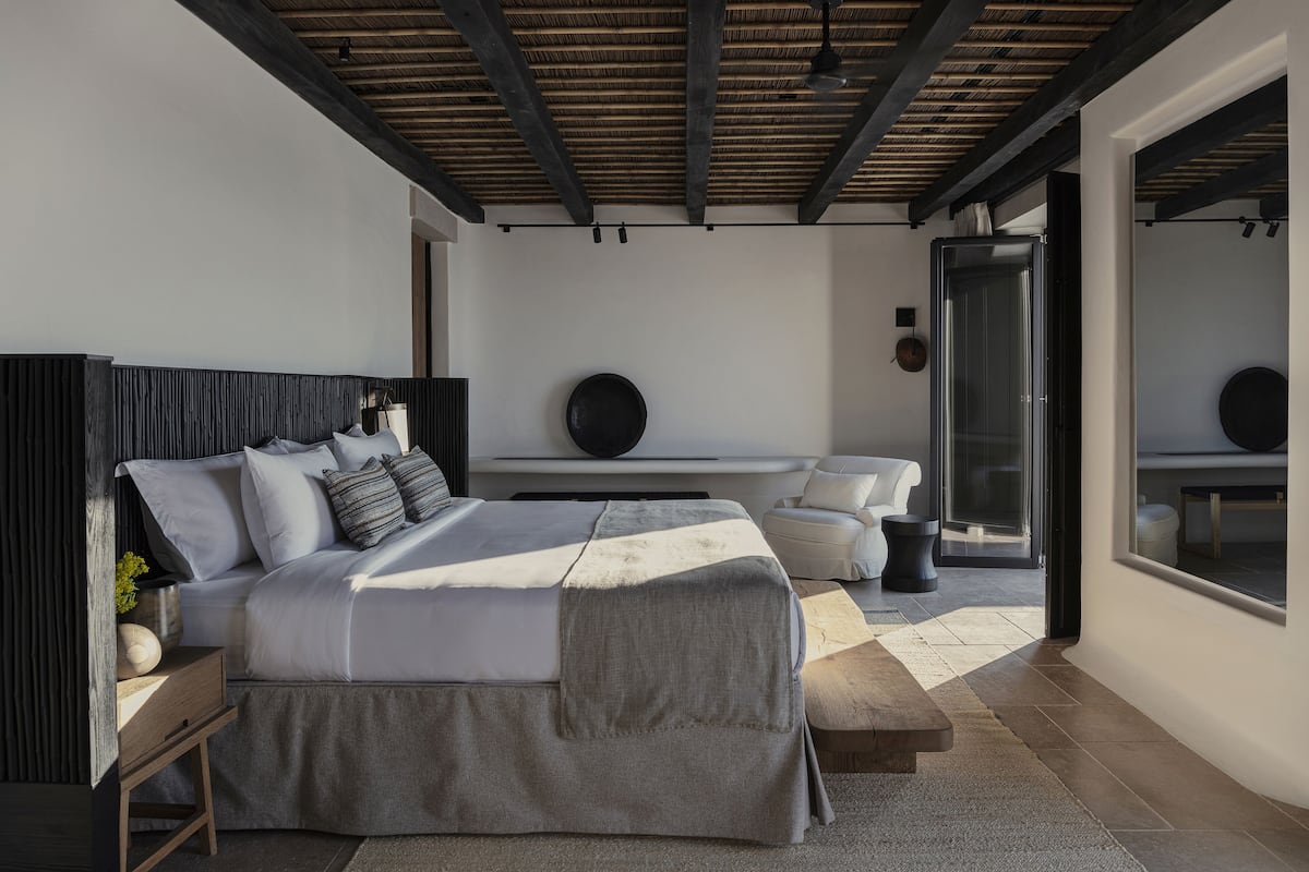 A modern bedroom with a large bed, a wooden headboard, a bedside table, a small stool, and a large window with a view of the outdoors.