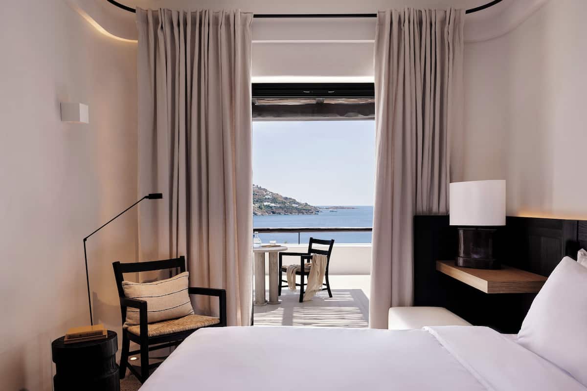 A hotel room with a bed, a chair, a table, and a view of the sea.