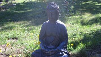 A Buddha statue in a garden setting with grass, flowers, and a house in the background.