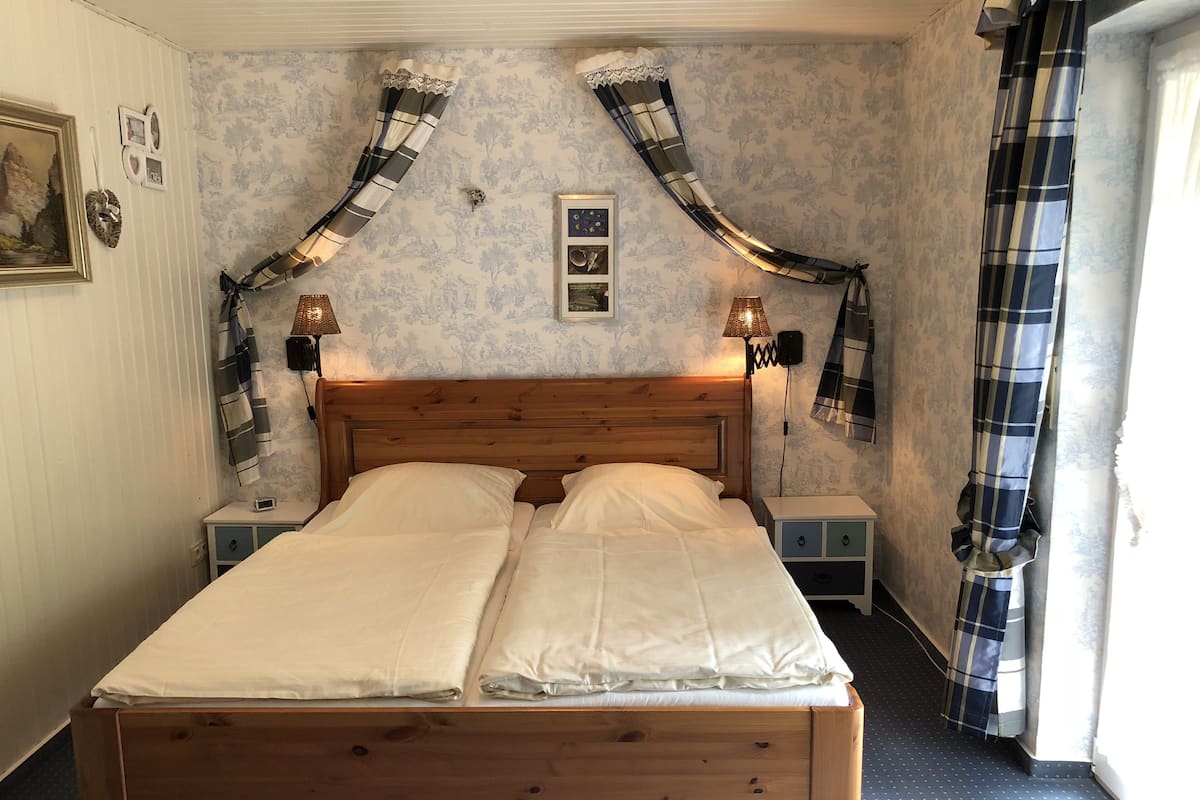 A double bed with wooden headboard, two bedside tables, a lamp, and a plaid curtain.