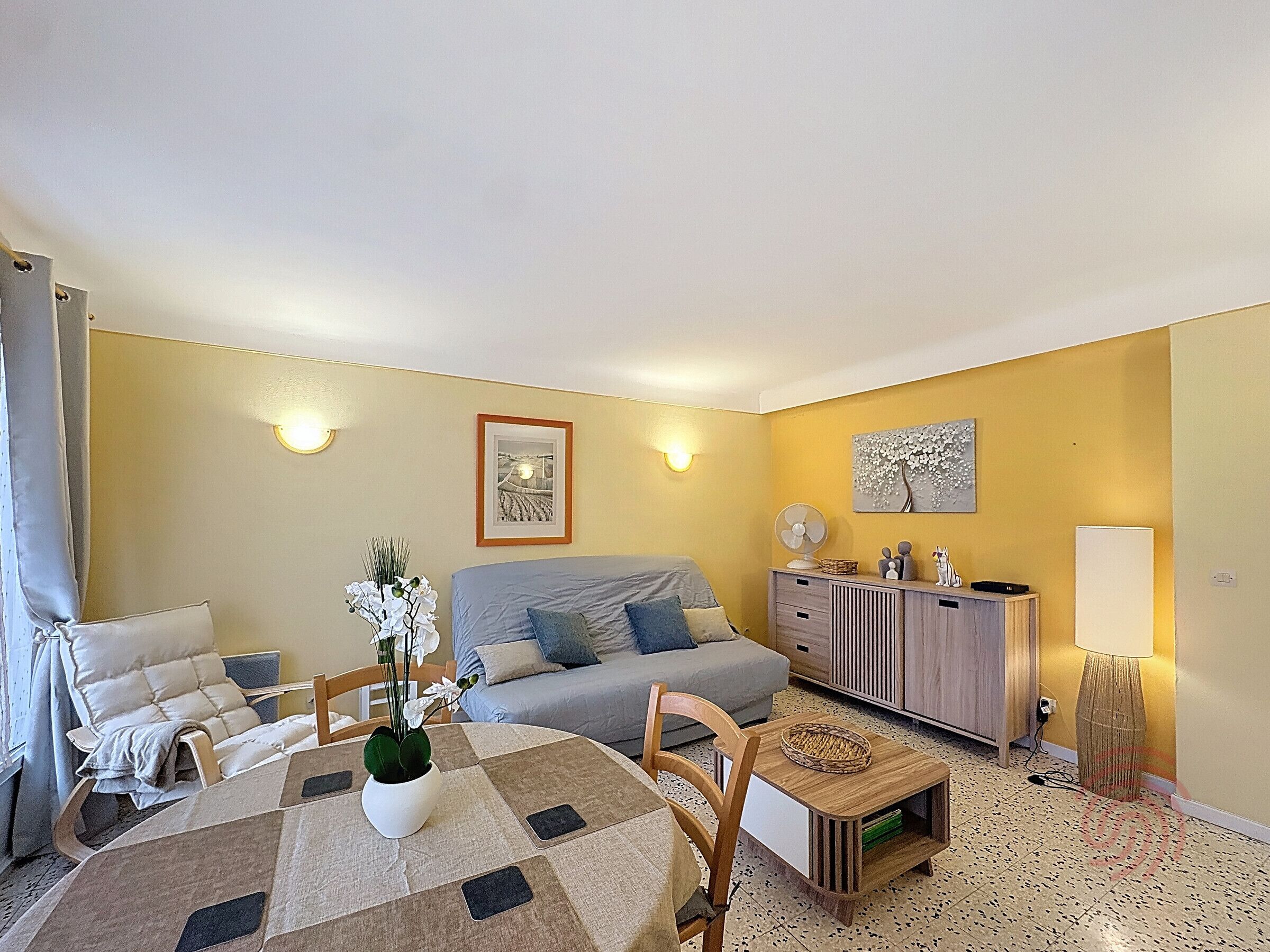 Een woonkamer met een bank, een eettafel met stoelen, een klein kastje en een lamp.