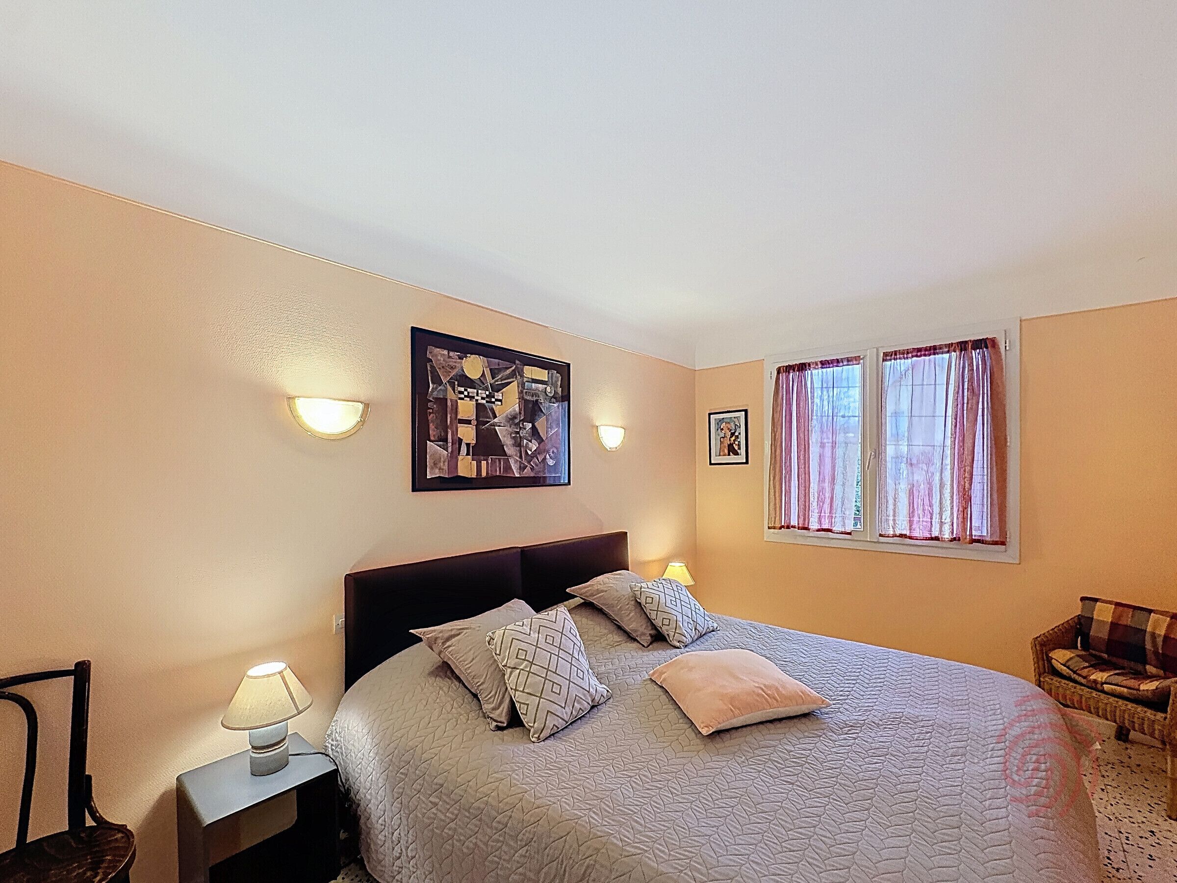 A bedroom with a bed, two bedside lamps, a wall art piece, a window with curtains, and a wicker chair.