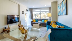 A modern living room with a sofa, a TV, a glass coffee table, and a view of buildings through large windows.