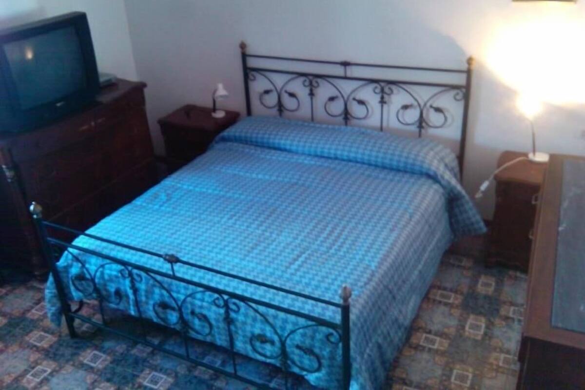 Une chambre à coucher avec un lit en métal, une couvre-lit à carreaux bleus, une petite table de chevet avec une lampe et un téléviseur posé sur une commode en bois.