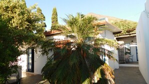 A courtyard with a palm tree, a white building, and a mountain in the background. - Case La Pergola (Santa Marina Salina)