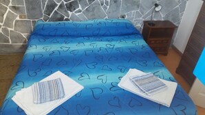 A bed with a blue heart-patterned bedspread, a wooden nightstand with a lamp, and a stone wall. - Case La Pergola (Santa Marina Salina)