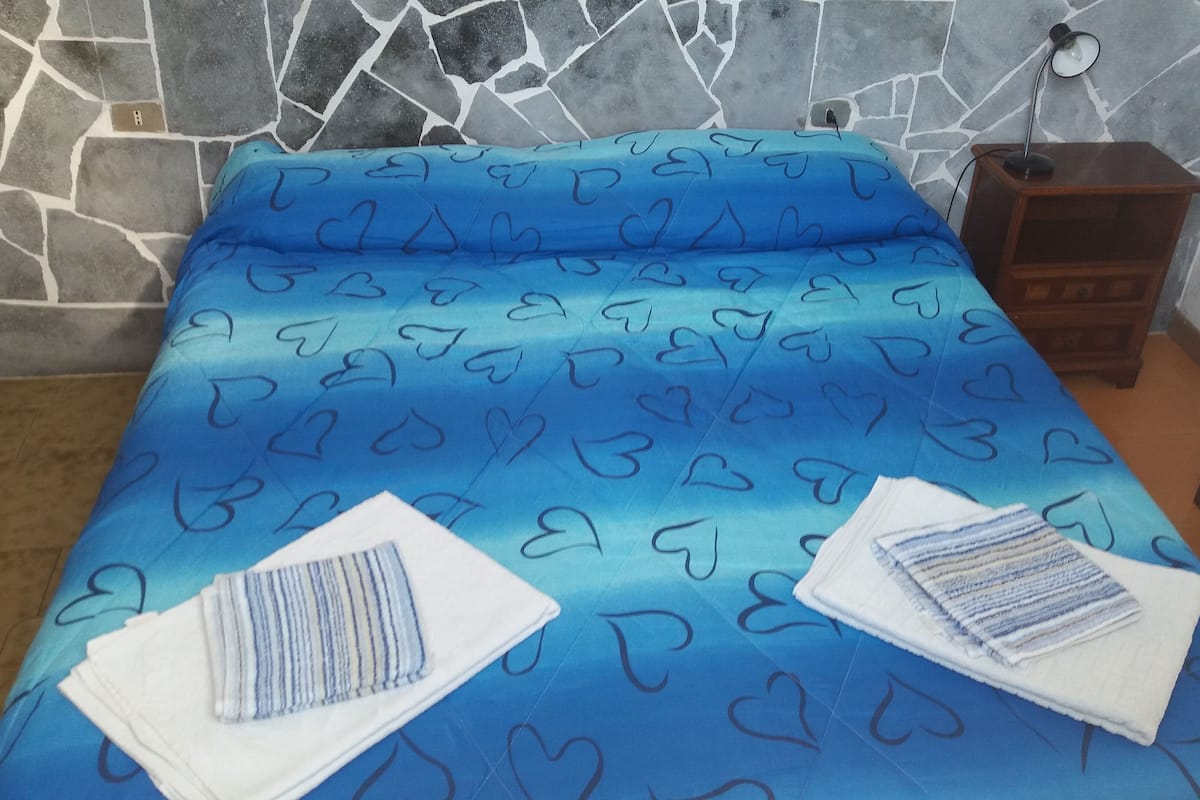 Un lit recouvert d’une couvre-lit à motifs de cœurs bleus, une table de chevet en bois avec une lampe, et un mur en pierre.