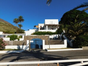 A modern building with a prominent blue gate, surrounded by greenery and palm trees. - Case La Pergola (Santa Marina Salina)