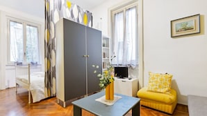 A room with a bed, a grey wardrobe, a small table with a vase, a yellow sofa, and a desk with a computer. - GuestHost - APT X3 Loreto Città Studi M1/M2 & C.so Buenos Aires (Milano)