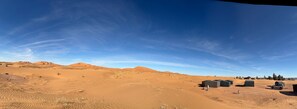 A desert landscape with sand dunes, a clear sky, and a few structures that appear to be part of a campsite or temporary accommodation. - Hoshizora sabaku camp (Rissani)