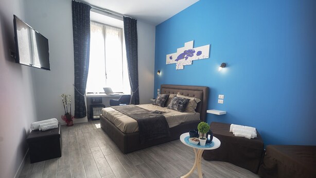 A modern bedroom with a large bed, a desk with a laptop, a small round table with decorative items, and a blue accent wall. - Roma Centrale Fidelis (Rome)