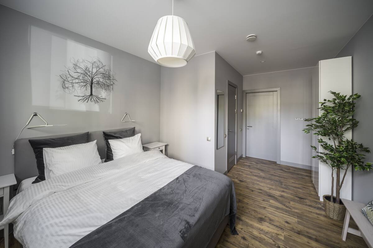 A modern bedroom with a large bed, bedside tables, a hanging light fixture, a wall-mounted artwork, and a wooden floor.