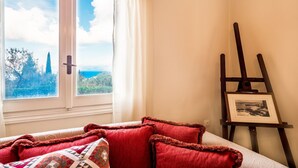 Une chambre avec un lit, une fenêtre donnant sur des arbres et des montagnes, et un tableau encadré posé sur un support.