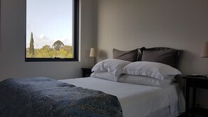 A bedroom with a large window, a bed with white and brown bedding, a bedside table with a lamp, and a view of greenery outside. - Stylish Parkside Apartment 2 Bed 2 Bath NETFLIX  Car park 12 minutes to City (Caulfield North)