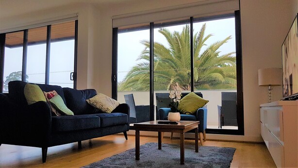A living room with a dark blue sofa, a small coffee table, and a view of palm trees through large windows. - Stylish Parkside Apartment 2 Bed 2 Bath NETFLIX  Car park 12 minutes to City (Caulfield North)