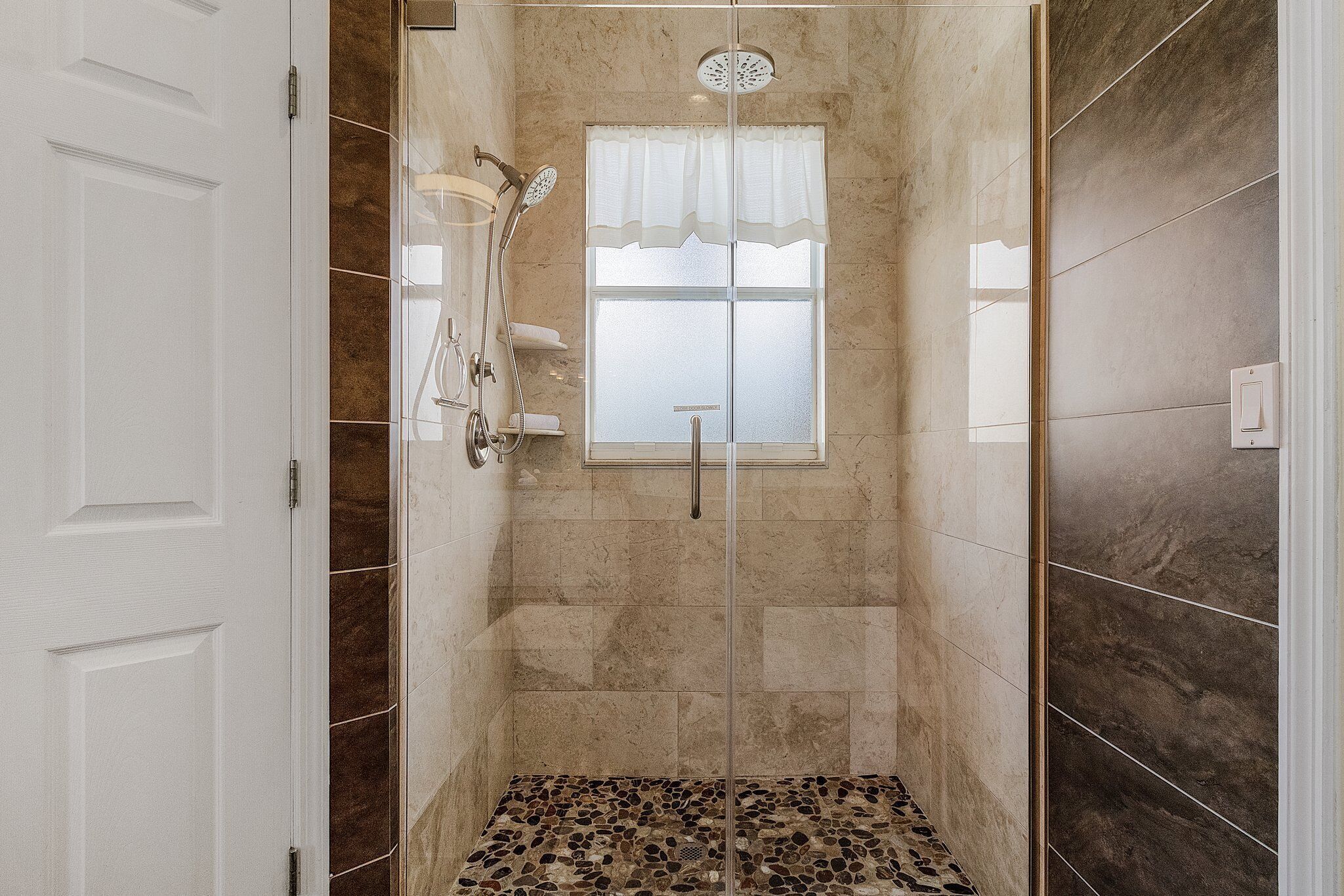 A walk-in shower with a glass door, a window with a white curtain, and a tiled floor.