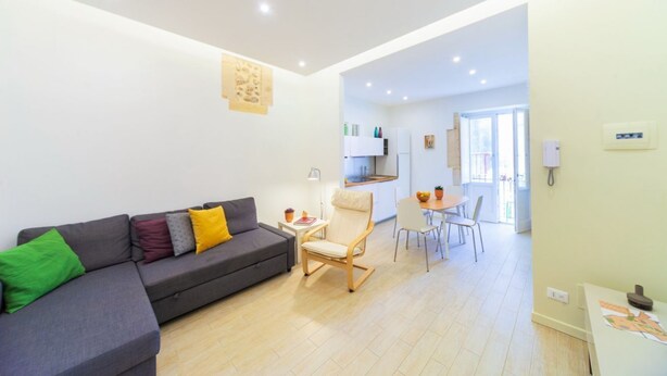 A modern living room with a grey sofa, a wooden chair, a dining table with chairs, and a kitchen in the background.