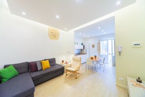 Un salon moderne avec un canapé gris, une chaise en bois, une table à manger avec des chaises, et une cuisine en arrière-plan.