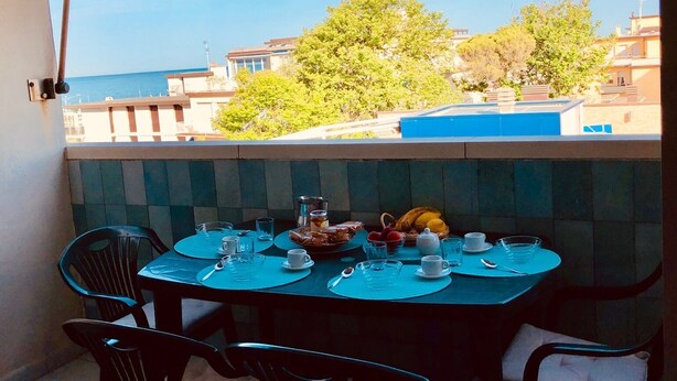 A balcony table set with plates, cups, and a fruit basket, overlooking a pool and buildings.