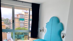 A bedroom with a large bed, a view of the city through a window, and a small wooden side table.