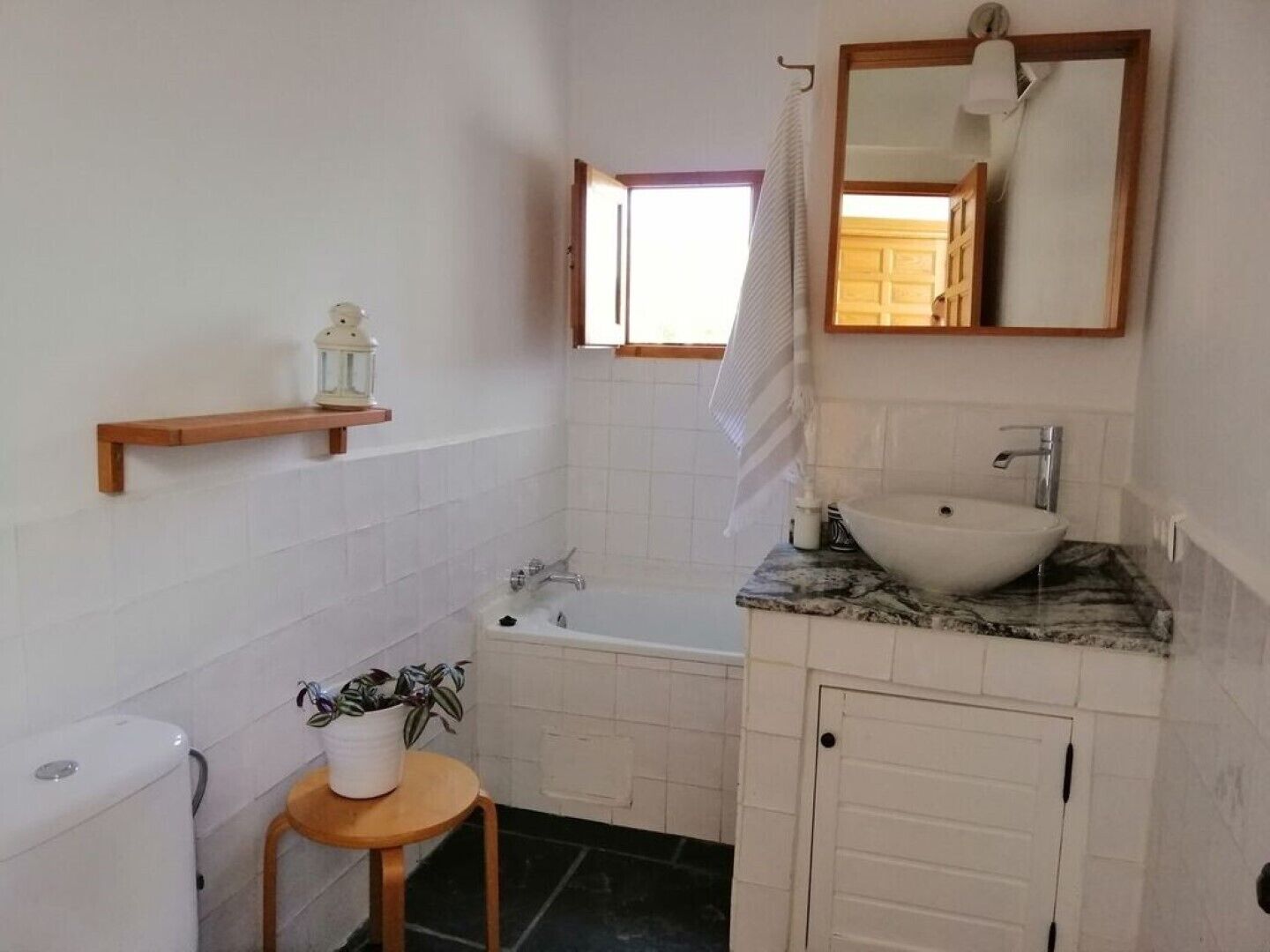 Een badkamer met een wit wastafel, een bad, een houten plank, een klein tafeltje met een potplant en een spiegel boven de wastafel.