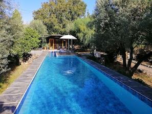 A rectangular swimming pool with a tiled edge, surrounded by trees and a covered seating area with chairs and a table. - Villa with Private Pool, Surrounded by Mountains, Olive, Walnut and Orange Trees (Orgiva)
