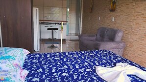 Un dormitorio con cama, una silla, una mesita y una kitchenette al fondo.