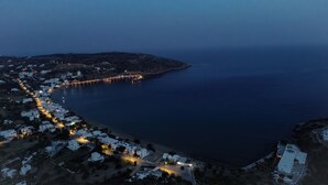 An aerial view of a coastal town at dusk with a curved road leading to the sea, illuminated buildings, and a bridge connecting the land to the island. - Evdokia Studios (Sifnos)