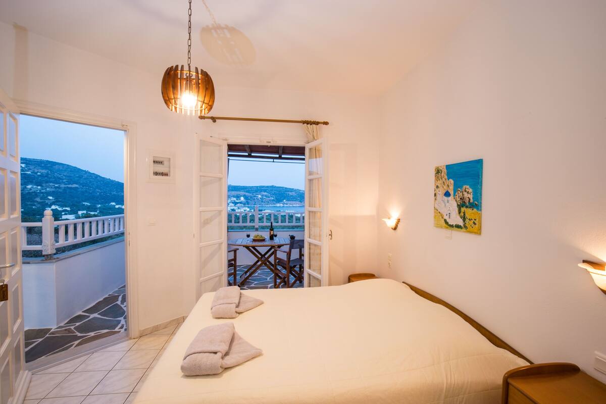 A hotel room with a bed, a table with chairs, a view of the sea, and a painting on the wall.