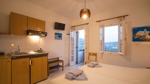 A compact hotel room with a kitchenette, a bed with towels, a balcony with a view, and a painting on the wall. - Evdokia Studios (Sifnos)