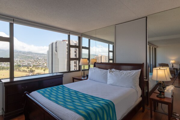 Habitación de hotel con una cama grande, una mesita de noche con lámpara, cómoda y vistas a un paisaje urbano a través de amplias ventanas.