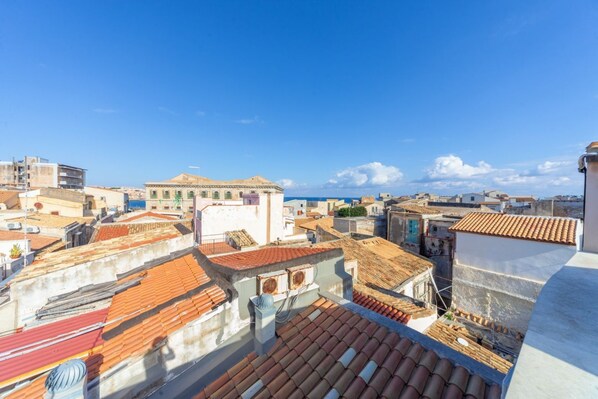A cityscape with terracotta-roofed buildings, a clear blue sky, and a distant view of the sea.