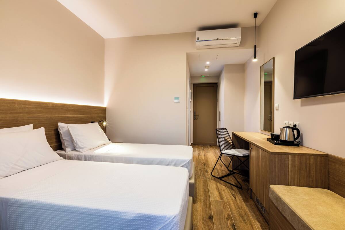 A hotel room with two beds, a desk with a chair, a TV, and a kitchenette area.