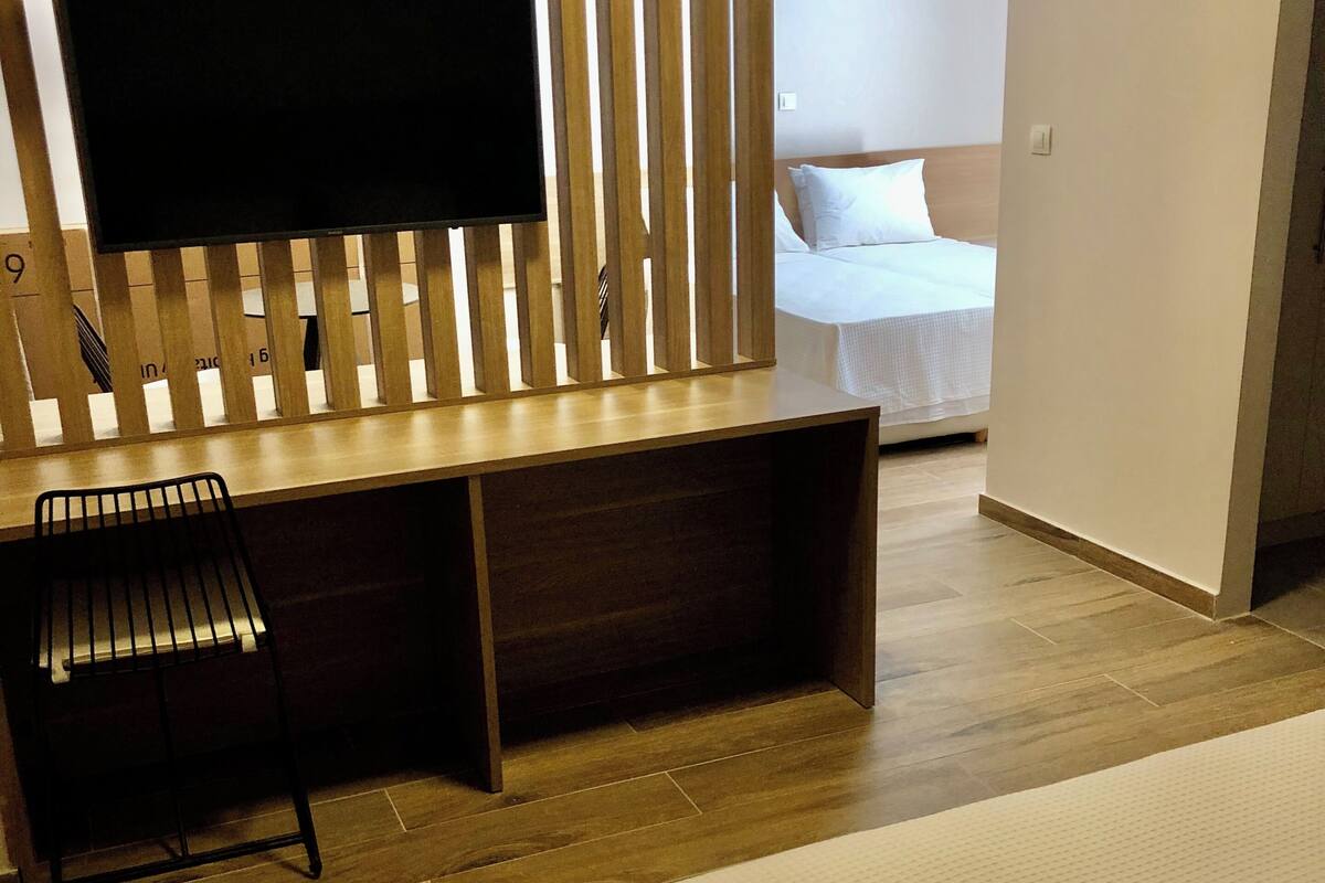 A hotel room with a bed, a desk with a chair, a TV mounted on a wooden slat wall, and a visible door leading to another room.