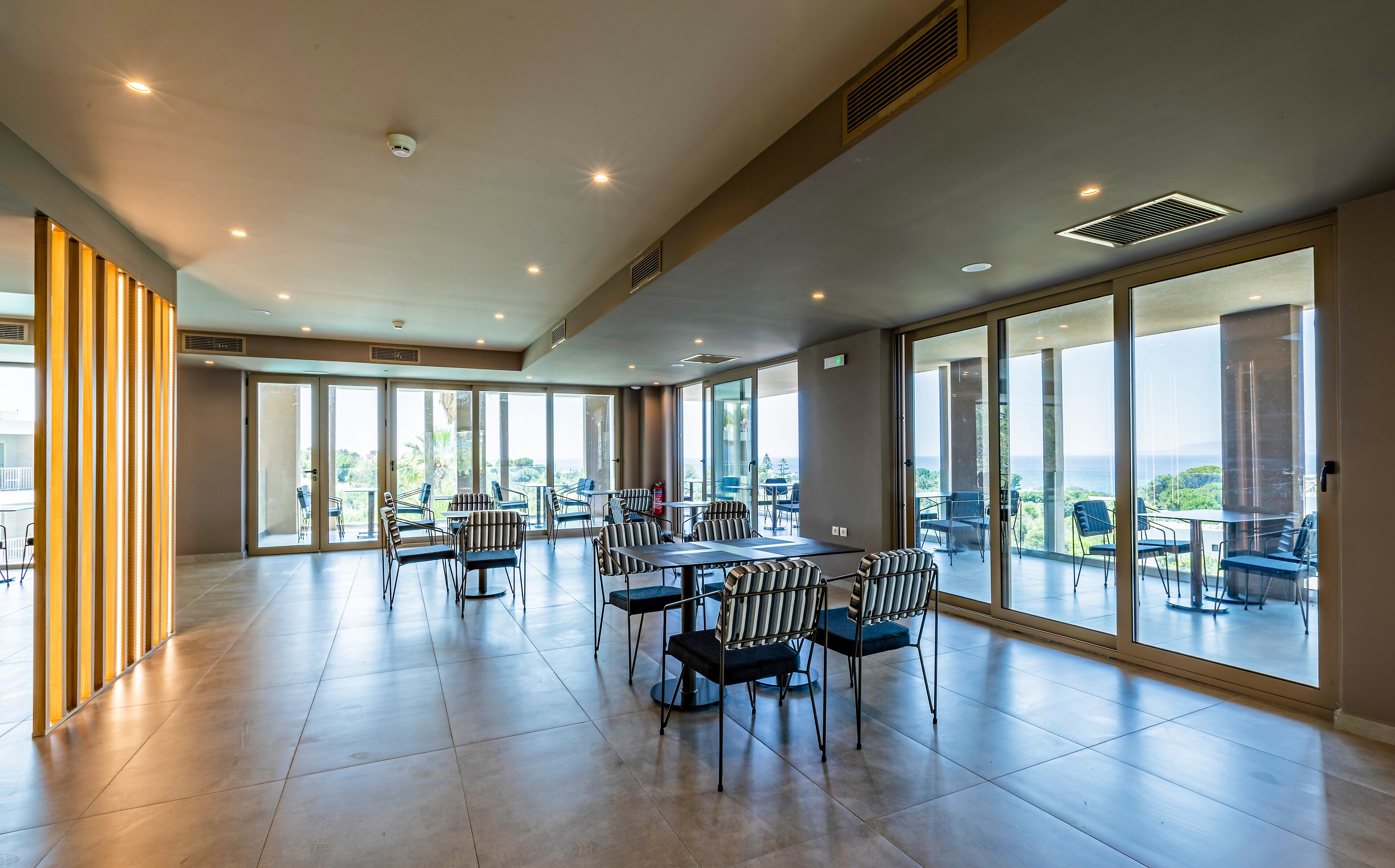 A spacious indoor area with a large table and chairs, large windows offering a view of the outdoors, and a fire extinguisher visible on the wall.