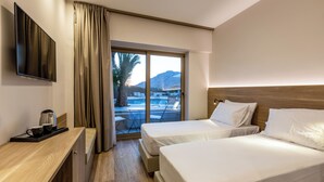 A hotel room with two beds, a TV, a desk with a coffee maker, and a view of a pool and mountains. - White Olive Premium Lindos - All Inclusive (Rhodes)