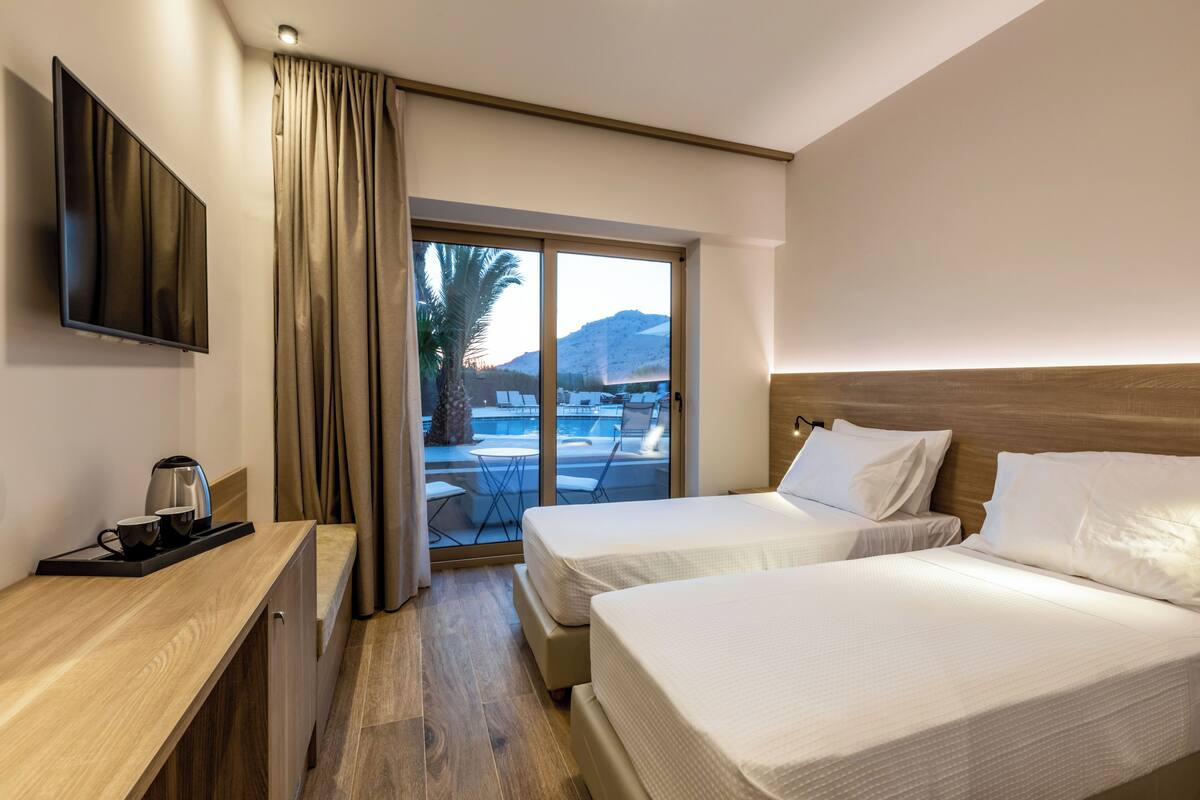 A hotel room with two beds, a TV, a desk with a coffee maker, and a view of a pool and mountains.