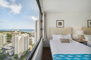 A hotel room with a large window, a bed with white bedding, two bedside tables with lamps, and framed pictures on the wall. - Block to Beach! Koko Resorts at the Waikiki Banyan 30th Floor Deluxe Ocean View,Free parking & Wifi (Honolulu)